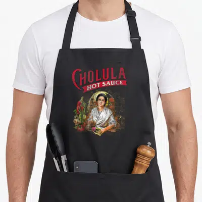 Cholula spice apron - Realistic woman under archway with hanging peppers and garlic below red Cholula Hot Sauce text — shown on a black cotton canvas apron against a white background