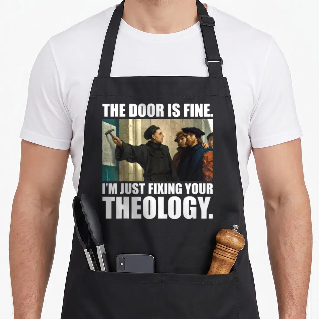 Martin meme apron - Man nailing paper to teal door with text The Door Is Fine I'm Just Fixing Your Theology — shown on a black cotton canvas apron against a white background