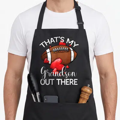 Funny grandson grandma aprons - Cartoon brown football with red hearts and white text reading That's My Grandson Out — shown on a black cotton canvas apron against a white background