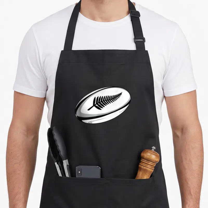 Jersey new zealand apron - White rugby ball with black fern leaf graphic on black bib apron — shown on a black cotton canvas apron against a white background