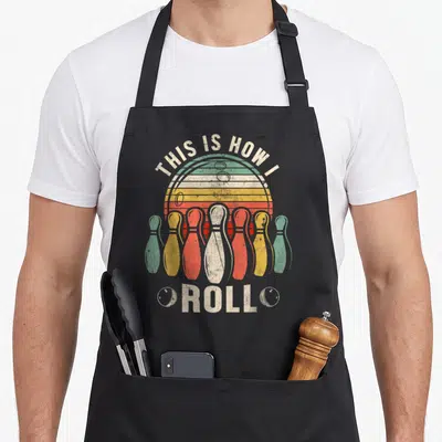 Bowler retro bowling apron - Seven bowling pins in front of a striped sun with text THIS IS HOW I ROLL — shown on a black cotton canvas apron against a white background