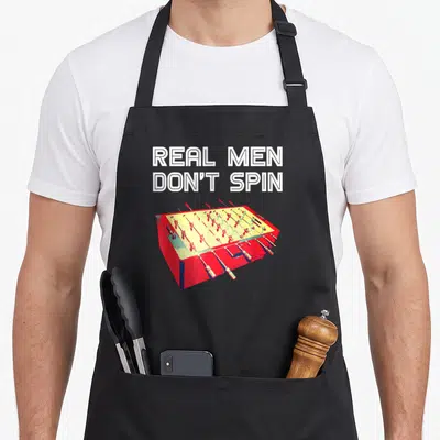Foosball spin apron - Real Men Don't Spin text above red yellow foosball table illustration — shown on a black cotton canvas apron against a white background