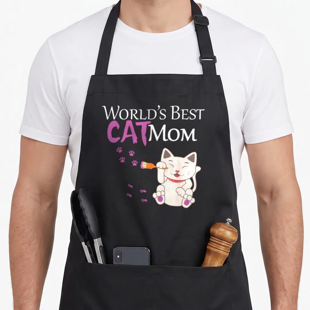 Funny cat lady apron - World's Best Cat Mom text above cartoon cat holding paintbrush with pink paw prints — shown on a black cotton canvas apron against a white background