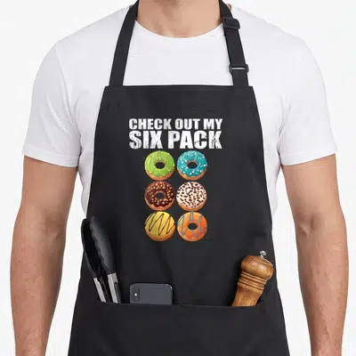 Check donut apron - CHECK OUT MY SIX PACK text above six colorful glazed donuts in two columns — shown on a black cotton canvas apron against a white background