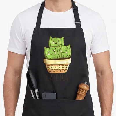 Catctus kitty cat apron - Three green cactus cats with pink cheeks in a tan pot above brown CATCTUS text — shown on a black cotton canvas apron against a white background