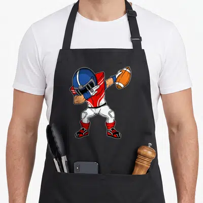 Dabbing football apron - Cartoon football player in red and blue uniform dabbing while holding an orange ball — shown on a black cotton canvas apron against a white background