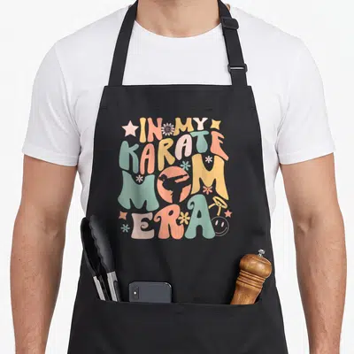 Karate mother's apron - Retro IN MY KARATE MOM ERA text with silhouette in O and colorful stars and flowers — shown on a black cotton canvas apron against a white background