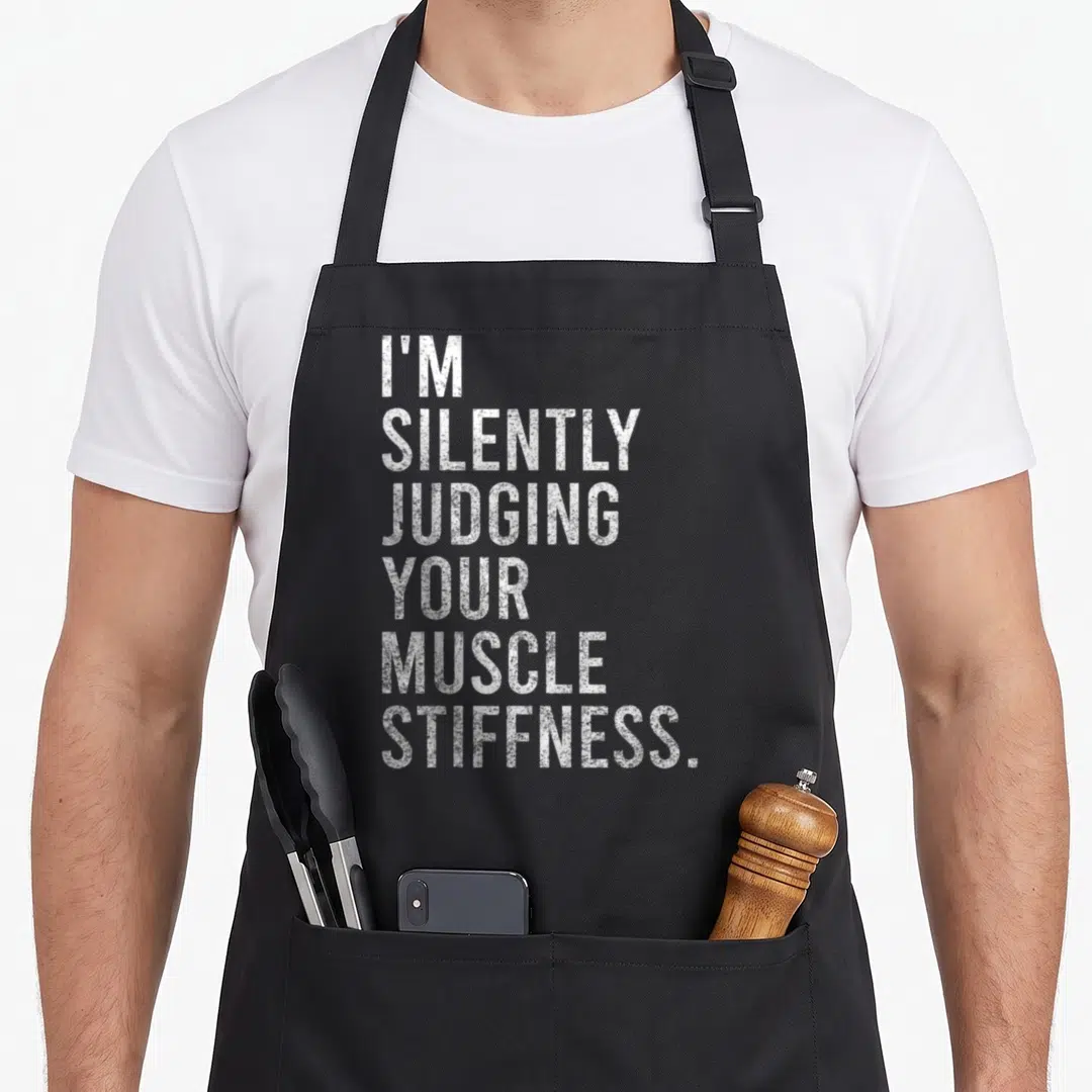 Silently massage therapist apron - White stacked text reading I'M SILENTLY JUDGING YOUR MUSCLE STIFFNESS on a dark — shown on a black cotton canvas apron against a white background