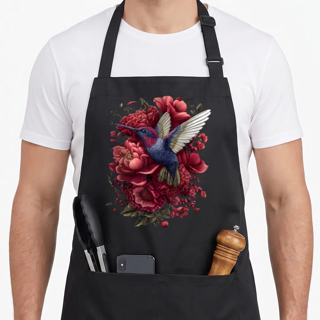 Enchanted the floral apron - Realistic blue hummingbird hovering over red flowers and gold leaves — shown on a black cotton canvas apron against a white background
