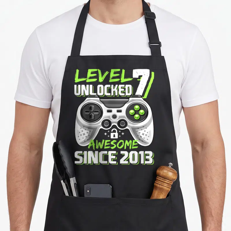 7th video game apron - Level 7 Unlocked controller graphic with Awesome Since 2013 text — shown on a black cotton canvas apron against a white background