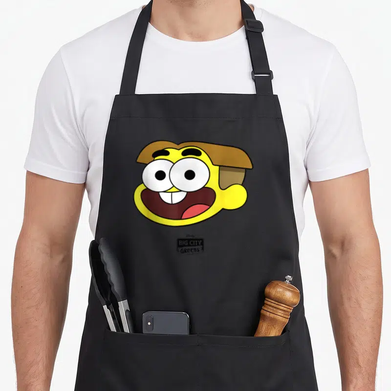 Funny channel cricket apron - Yellow cartoon character head with brown hair and BIG CITY Greens logo below — shown on a black cotton canvas apron against a white background
