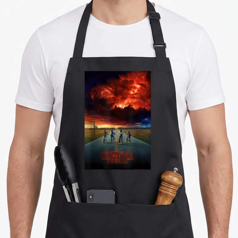 Demogorgon stranger apron - Four boys on bikes under red stormy sky with Welcome to Hawkins sign — shown on a black cotton canvas apron against a white background