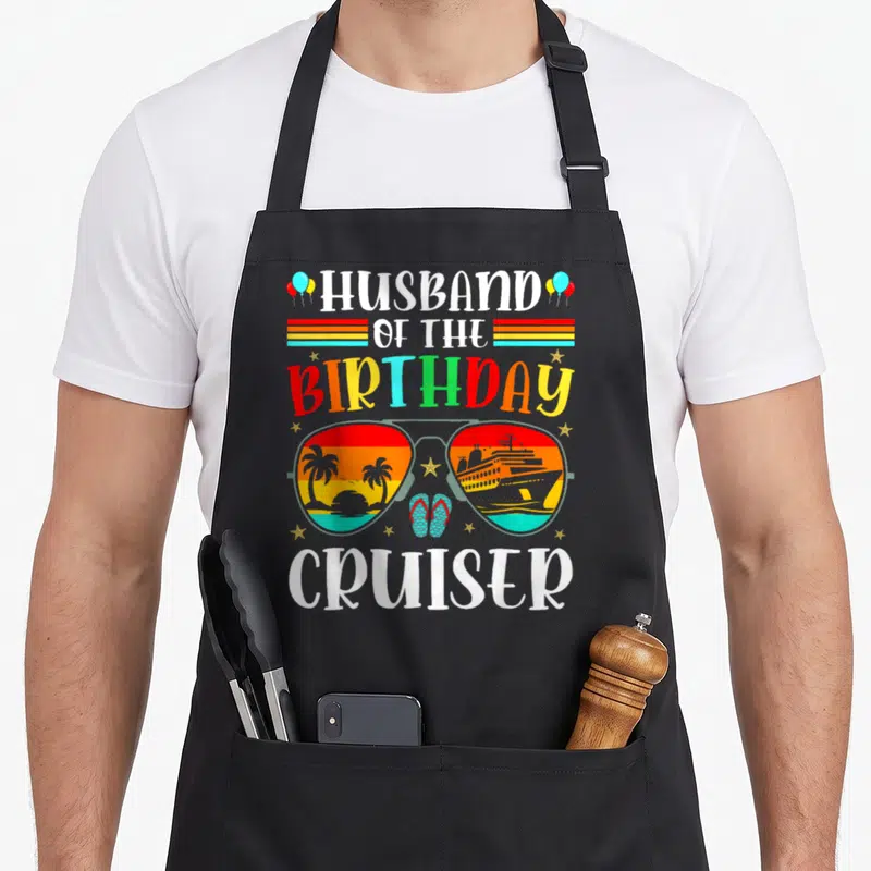 Cruiser husband apron - Sunglasses with beach and ship lenses, text Husband of the Birthday Cruiser — shown on a black cotton canvas apron against a white background