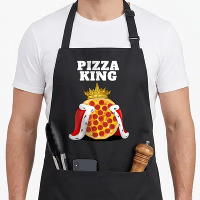 Italian pizza king apron - Cartoon pepperoni pizza wearing a gold crown and red cape below PIZZA KING text — shown on a black cotton canvas apron against a white background