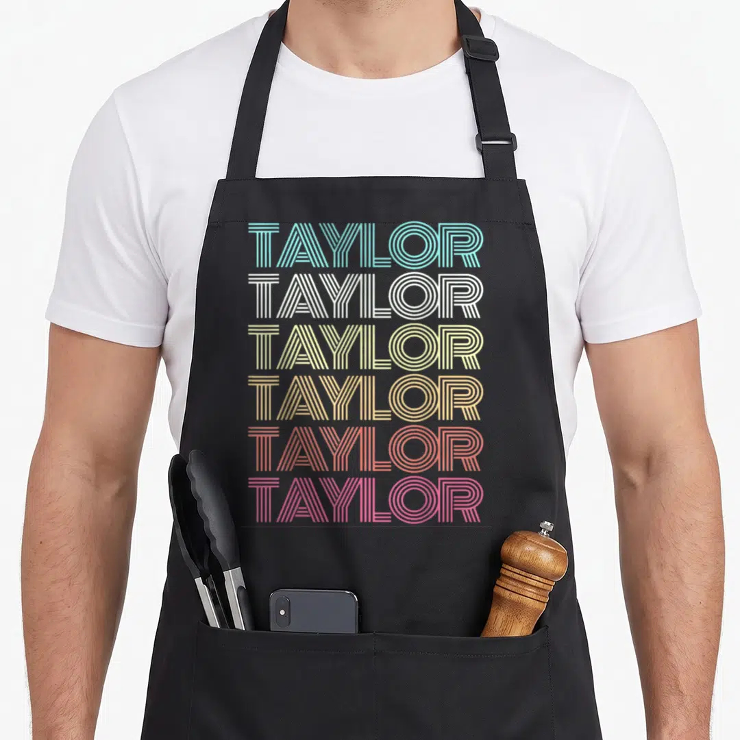 Taylor groovy 80s apron - Six stacked lines of TAYLOR text in blue white yellow orange red and pink linear font — shown on a black cotton canvas apron against a white background