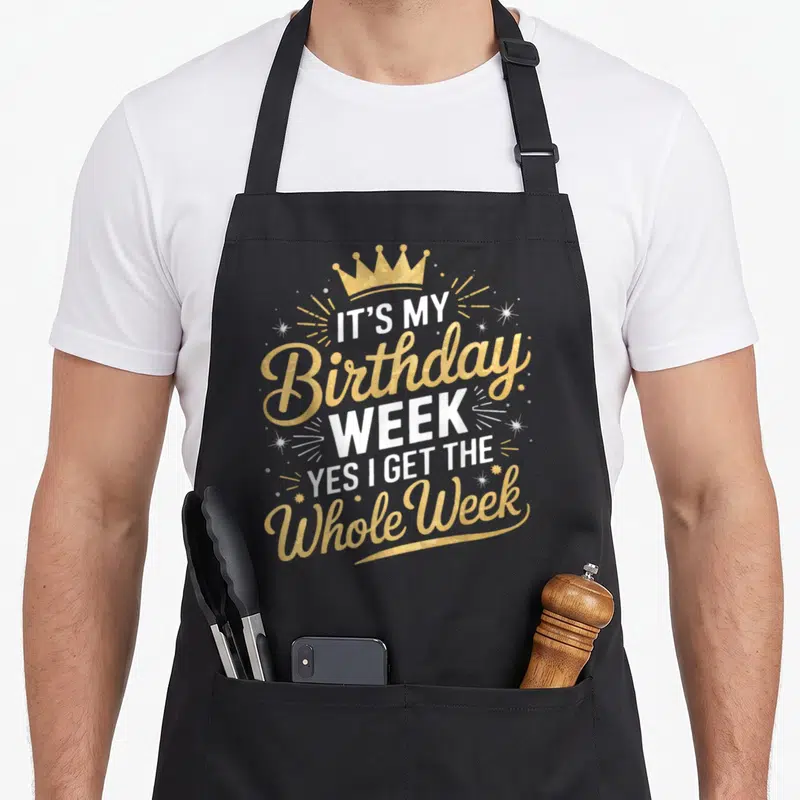 Cute whole birthday apron - Gold crown and white text reading It's My Birthday Week Yes I Get The Whole Week — shown on a black cotton canvas apron against a white background