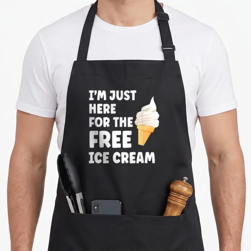 Free ice cream apron - Stacked white text reading I'M JUST HERE FOR THE FREE ICE CREAM next to orange cone with white — shown on a black cotton canvas apron against a white background