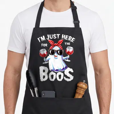 Boo ghost halloween apron - Cartoon ghost in sunglasses holding two red wine glasses under arched text reading I'M JUST — shown on a black cotton canvas apron against a white background