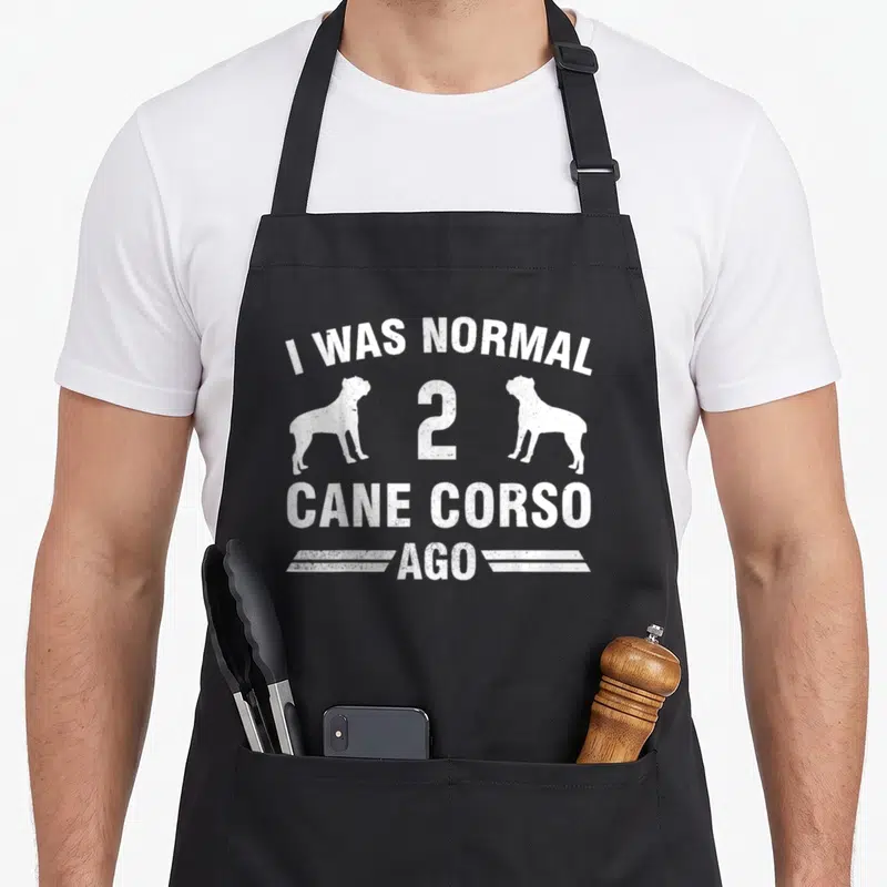 Normal cane apron - White distressed text I Was Normal 2 Cane Corso Ago with two dog silhouettes — shown on a black cotton canvas apron against a white background