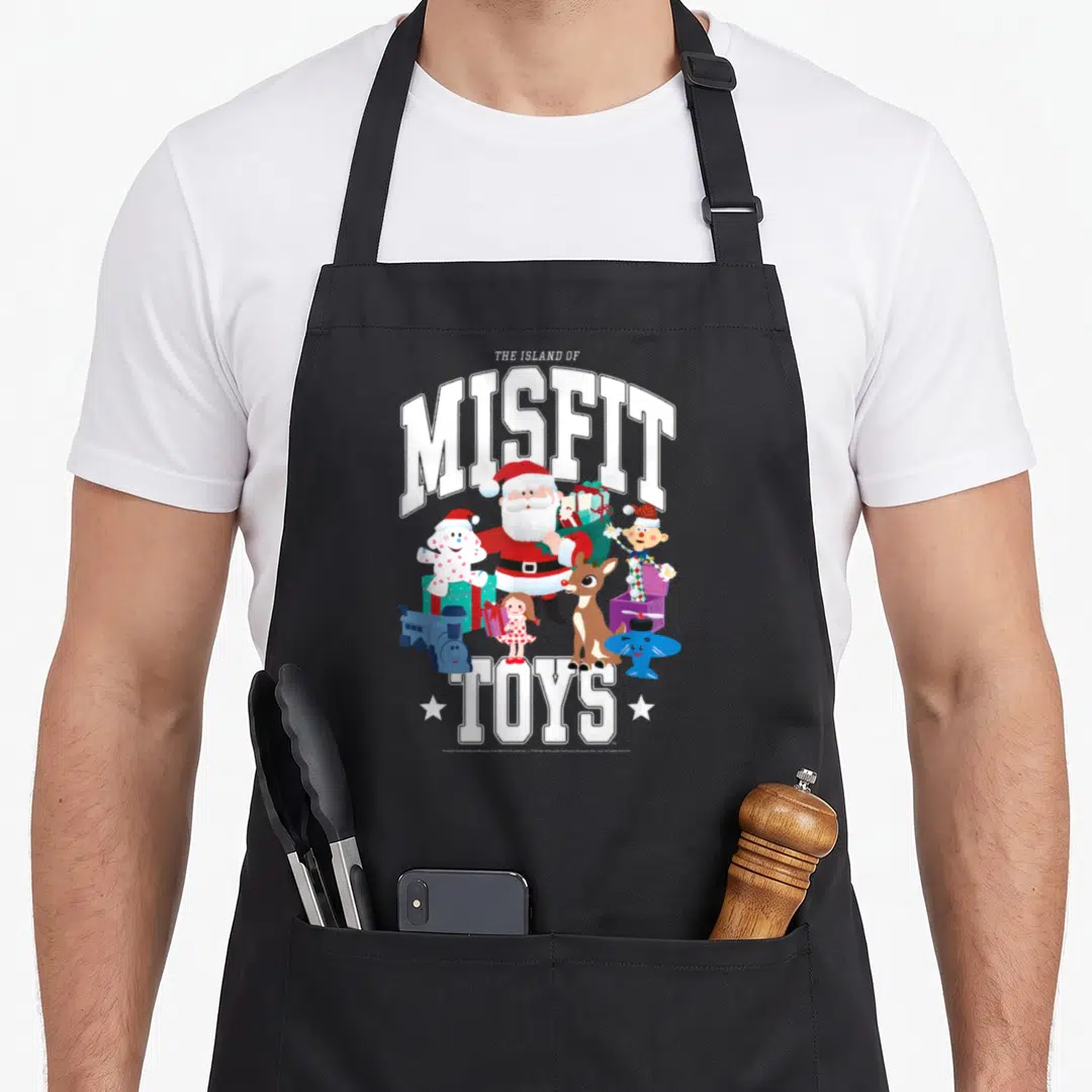 Rudolph christmas apron - Santa Claus and toys under The Island of Misfit Toys varsity text in white, red, blue — shown on a black cotton canvas apron against a white background