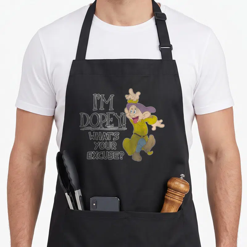 Excuse snow white apron - I'M DOPEY! WHAT'S YOUR EXCUSE? text with standing cartoon character in olive green and purple — shown on a black cotton canvas apron against a white background