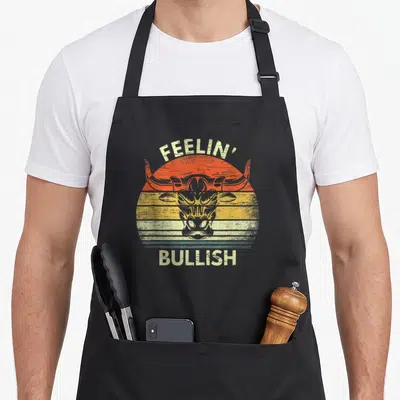 Feelin stock aprons - Retro bull head with FEELIN' BULLISH text over striped orange yellow grey sun — shown on a black cotton canvas apron against a white background