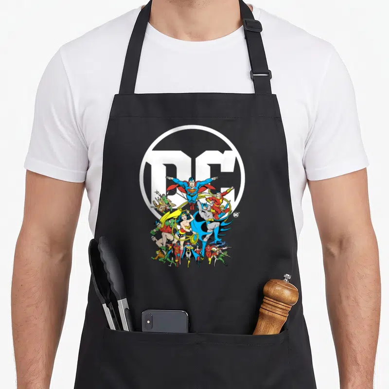 Poster character aprons - Superheroes clustered in front of a white DC logo circle in cartoon style — shown on a black cotton canvas apron against a white background