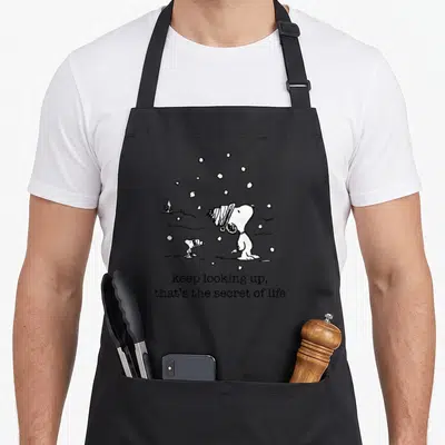 Woodstock christmas apron - Snoopy and Woodstock looking up at snowflakes with text keep looking up that’s the secret — shown on a black cotton canvas apron against a white background