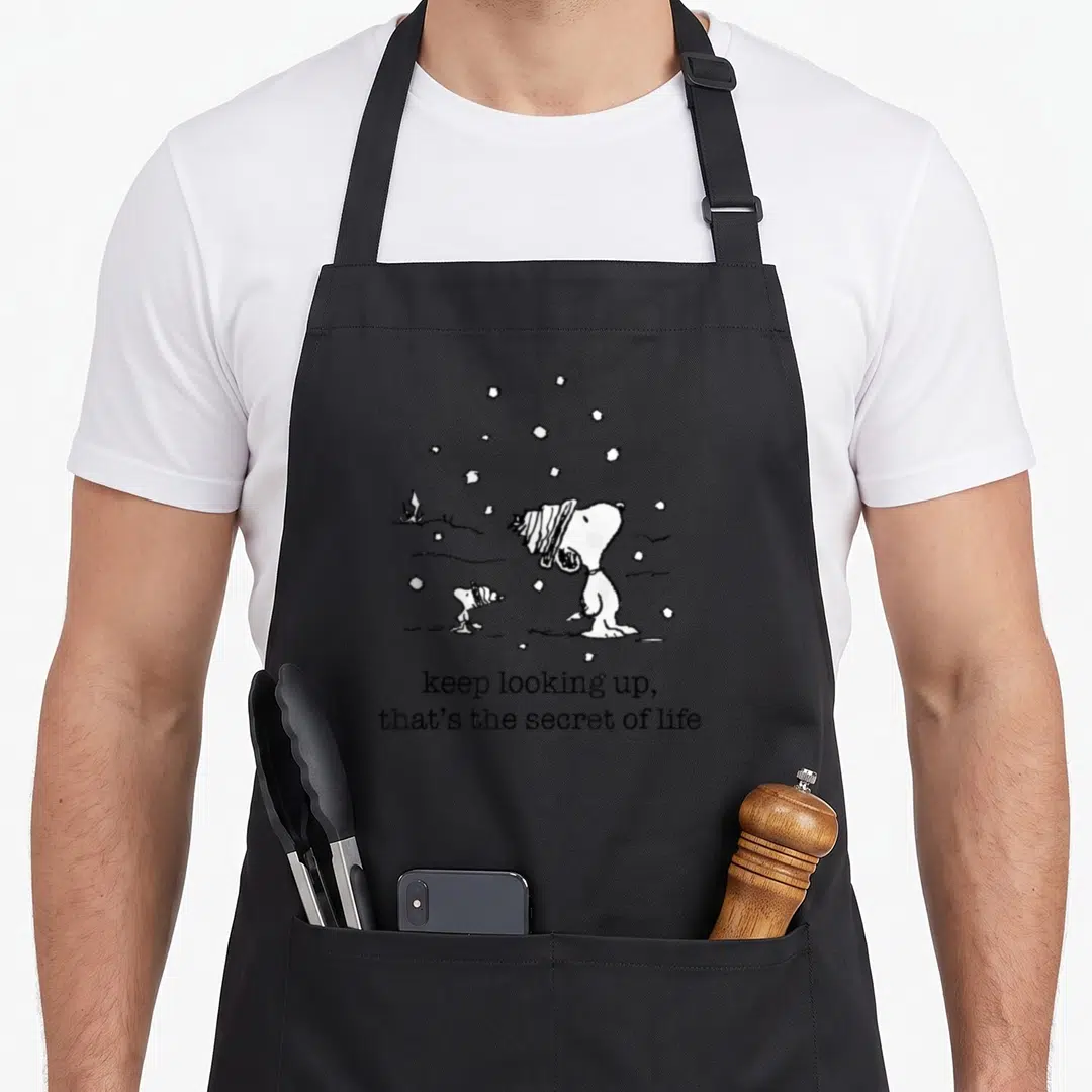 Woodstock christmas apron - Snoopy and Woodstock looking up at snowflakes with text keep looking up that’s the secret — shown on a black cotton canvas apron against a white background