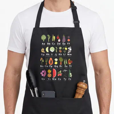 Garden veggie apron - Alphabet grid of 26 produce items including Ube, Ximmea, and Yarrow in cartoon style — shown on a black cotton canvas apron against a white background