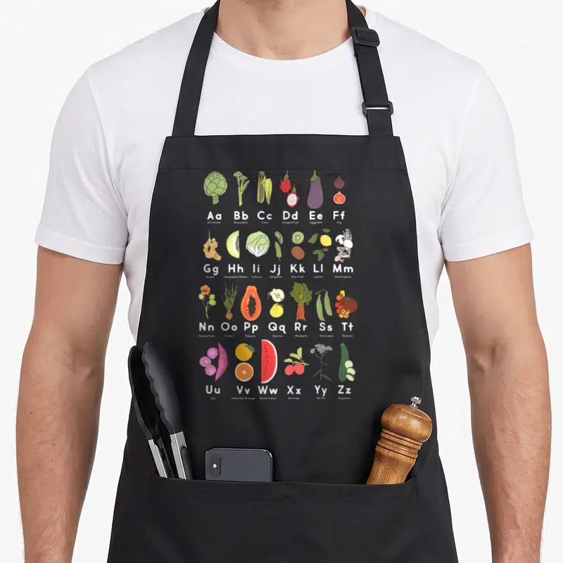 Garden veggie apron - Alphabet grid of 26 produce items including Ube, Ximmea, and Yarrow in cartoon style — shown on a black cotton canvas apron against a white background
