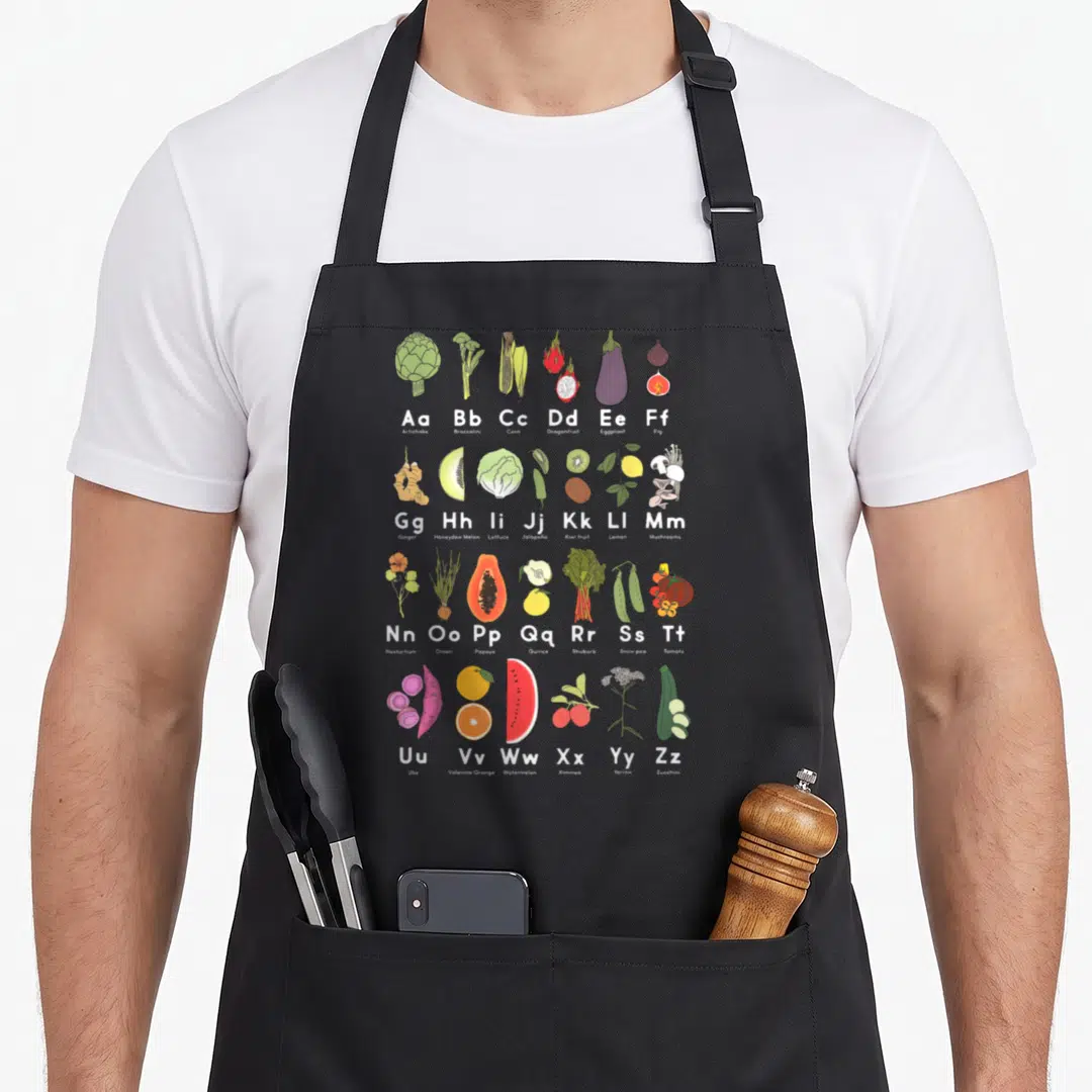 Garden veggie apron - Alphabet grid of 26 produce items including Ube, Ximmea, and Yarrow in cartoon style — shown on a black cotton canvas apron against a white background