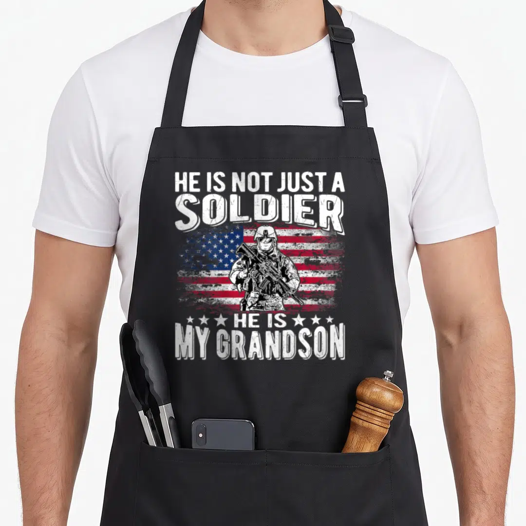 Grandson patriotic apron - Soldier holding rifle in front of distressed USA flag with white text HE IS NOT JUST — shown on a black cotton canvas apron against a white background