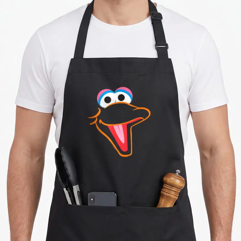 Face bird apron - Stylized cartoon bird face with orange beak, blue and pink eyes, and open mouth — shown on a black cotton canvas apron against a white background