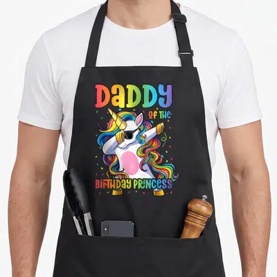 Princess daddy apron - Cartoon white unicorn dabbing with rainbow mane and DaDDY OF THE BIRTHDAY PRINCESS text — shown on a black cotton canvas apron against a white background