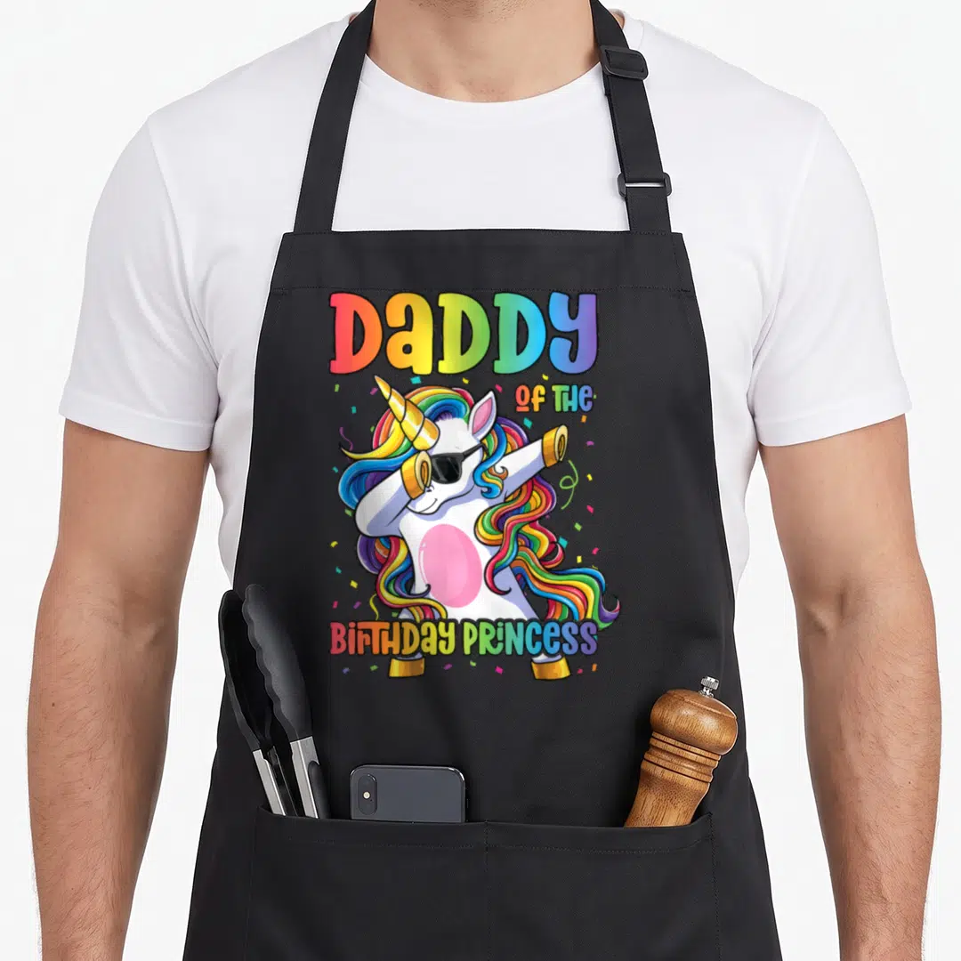 Princess daddy apron - Cartoon white unicorn dabbing with rainbow mane and DaDDY OF THE BIRTHDAY PRINCESS text — shown on a black cotton canvas apron against a white background