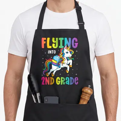 Second unicorn apron - Cartoon white unicorn with rainbow mane and backpack under Flying Into 2nd Grade text — shown on a black cotton canvas apron against a white background