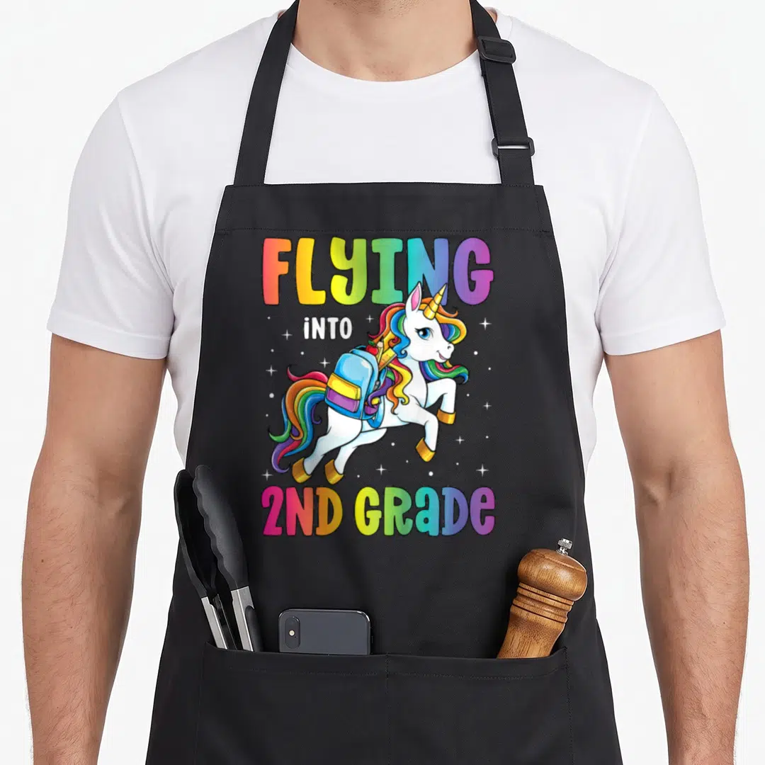 Second unicorn apron - Cartoon white unicorn with rainbow mane and backpack under Flying Into 2nd Grade text — shown on a black cotton canvas apron against a white background
