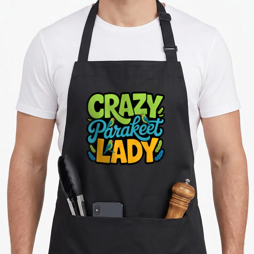 Obsessed bird apron - CRAZY Parakeet LADY text with three cartoon parakeets perched on lime, blue, and yellow letters — shown on a black cotton canvas apron against a white background