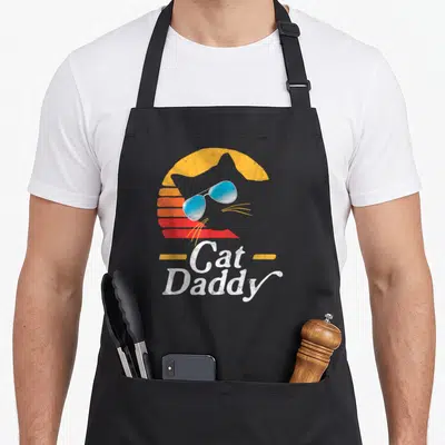 Daddy cat apron - Retro cat head silhouette with blue sunglasses and striped sun above Cat Daddy text — shown on a black cotton canvas apron against a white background