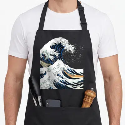 Kanagawa hokusai japanese wave apron - Cresting blue and white wave with two tan boats in ukiyo-e style — shown on a black cotton canvas apron against a white background