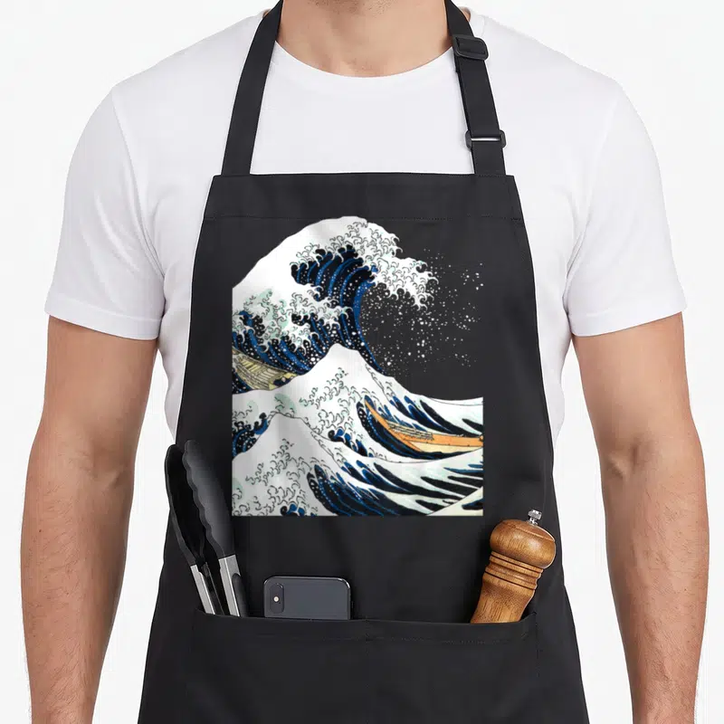 Kanagawa hokusai japanese wave apron - Cresting blue and white wave with two tan boats in ukiyo-e style — shown on a black cotton canvas apron against a white background