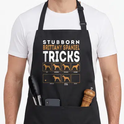Spaniel dog training apron - Tan dog silhouettes in boxes labeled Sit, Down, Shake, Come, Fetch, Roll Over, Stay — shown on a black cotton canvas apron against a white background