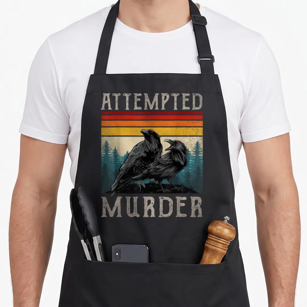 Attempted ravens apron - Two black ravens in front of teal trees under red orange yellow stripes with text — shown on a black cotton canvas apron against a white background