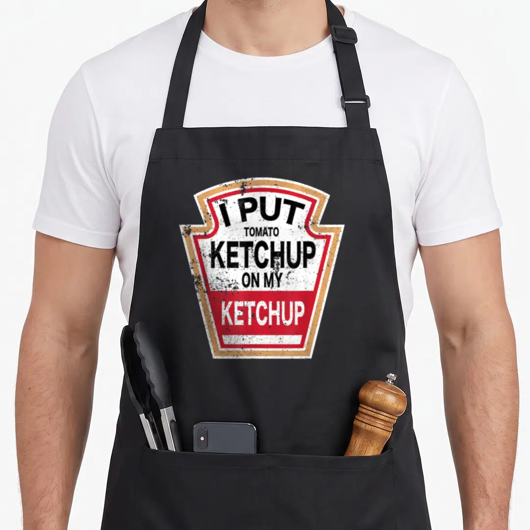 Tomato halloween apron - Shield sign graphic with stacked text I PUT TOMATO KETCHUP ON MY KETCHUP in distressed red — shown on a black cotton canvas apron against a white background