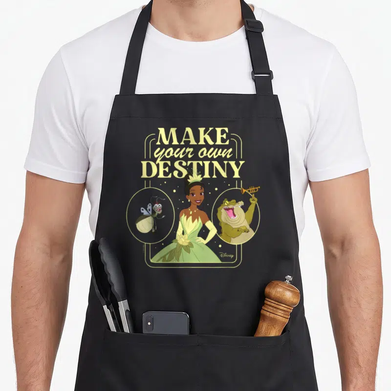 Tiana magic apron - Make your own destiny text above Tiana with frog and alligator in circles, yellow green brown — shown on a black cotton canvas apron against a white background