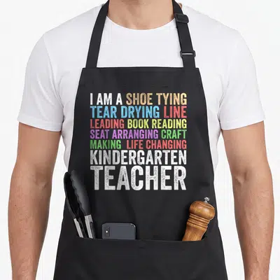 Kindergarten creative apron - Stacked rainbow text listing kindergarten teacher duties like shoe tying and book reading — shown on a black cotton canvas apron against a white background