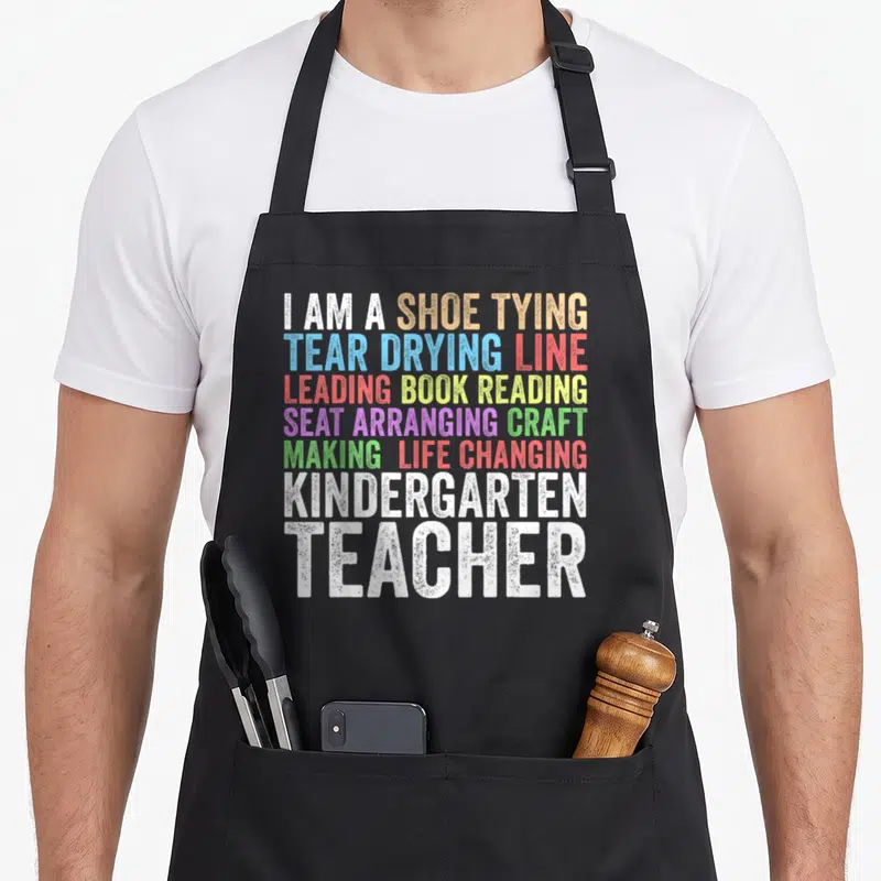 Kindergarten creative apron - Stacked rainbow text listing kindergarten teacher duties like shoe tying and book reading — shown on a black cotton canvas apron against a white background