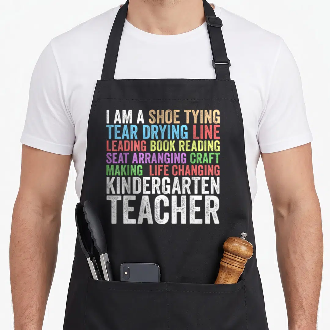 Kindergarten creative apron - Stacked rainbow text listing kindergarten teacher duties like shoe tying and book reading — shown on a black cotton canvas apron against a white background