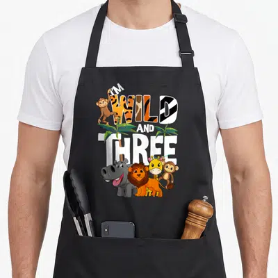 Wild safari birthday apron - I'M WILD AND THREE text with cartoon animals and palm trees in orange and white — shown on a black cotton canvas apron against a white background
